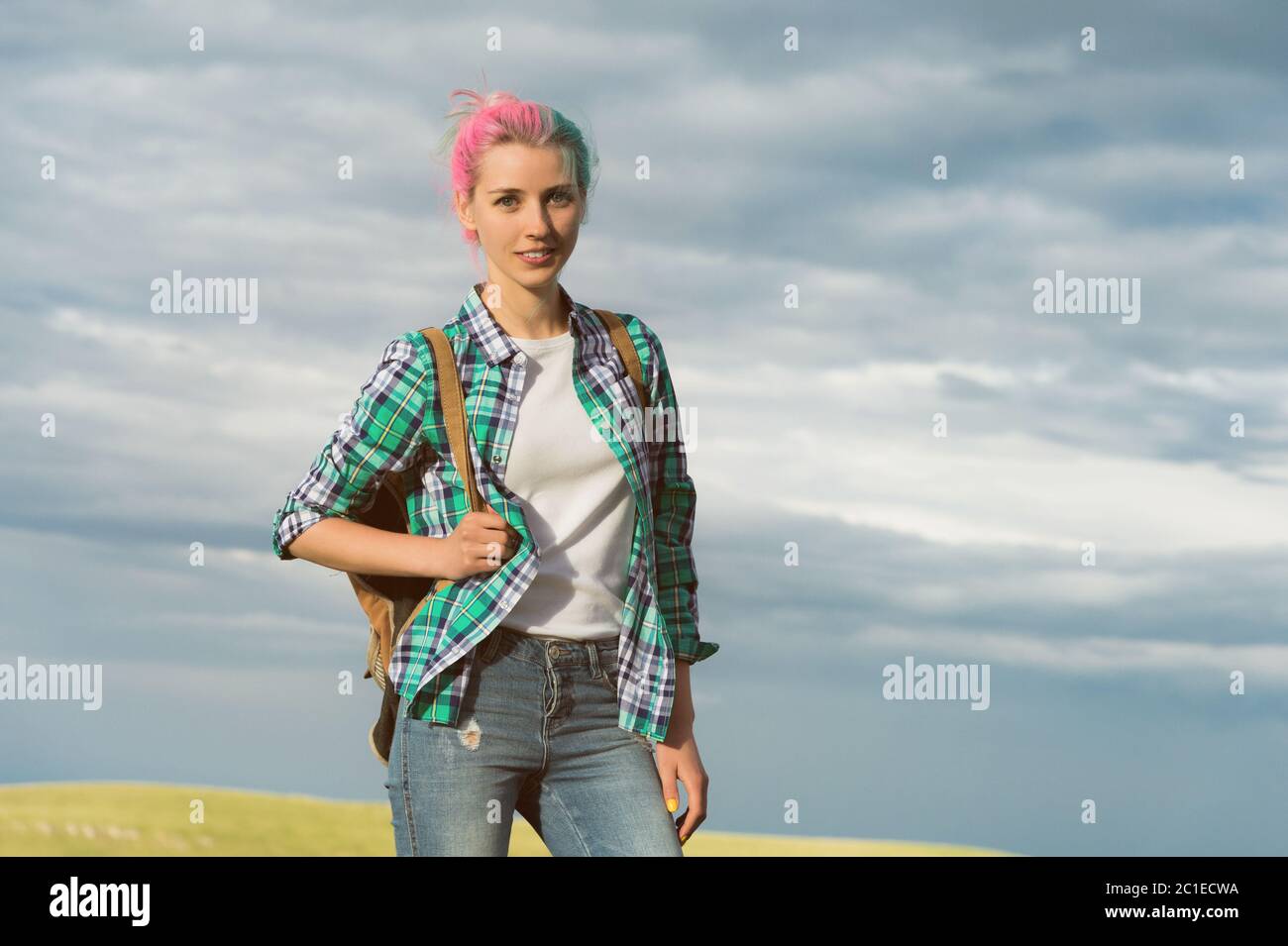 ritratto di una giovane donna sorridente attraente in abiti da camicia plaid con piccolo zaino in giornata di sole sul cielo nuvoloso e campo Foto Stock