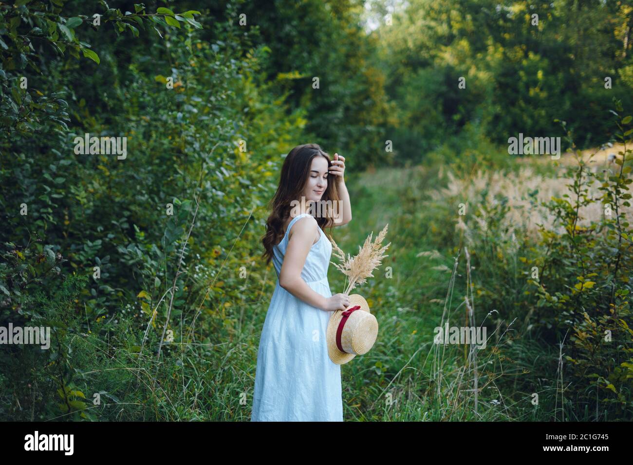 Bella ragazza in un abito bianco con un bouquet di fiori secchi. Giovane donna in un parco verde. Ragazza in un abito e una giornata di sole in estate Foto Stock