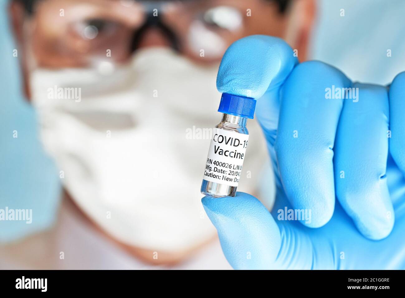 Mano in guanti medici blu che tengono il flaconcino piccolo con etichetta vaccino Coronavirus (proprio disegno adesivo, non prodotto reale), viso sfocato in maschera di cotone e. Foto Stock