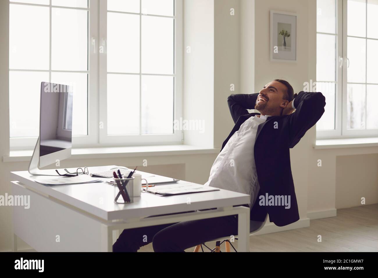 Uomo d'affari felice con le mani dietro la testa sorridente riposo corre sogni a una scrivania in ufficio. Foto Stock