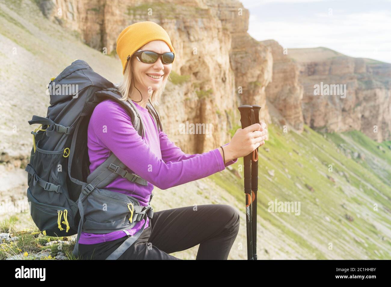 Sorridi la ragazza in un cappello giallo e un paio di occhiali da sole che si siede ai piedi di epiche rocce con uno zaino successivo e guarda Foto Stock