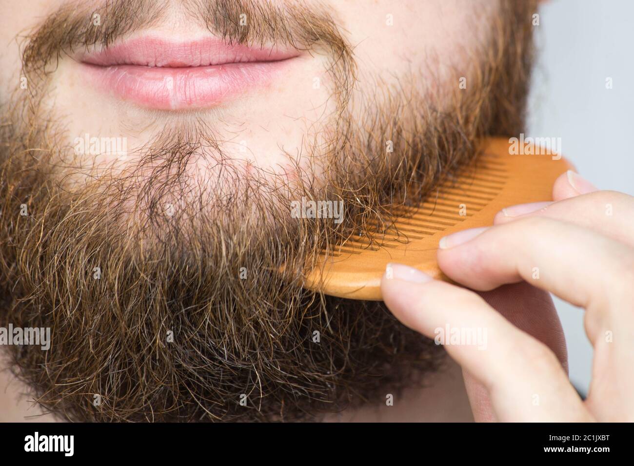 Primo piano di un giovane uomo lo styling della sua lunga barba con un pettine mentre sta in piedi da solo in un monolocale contro uno sfondo bianco Foto Stock