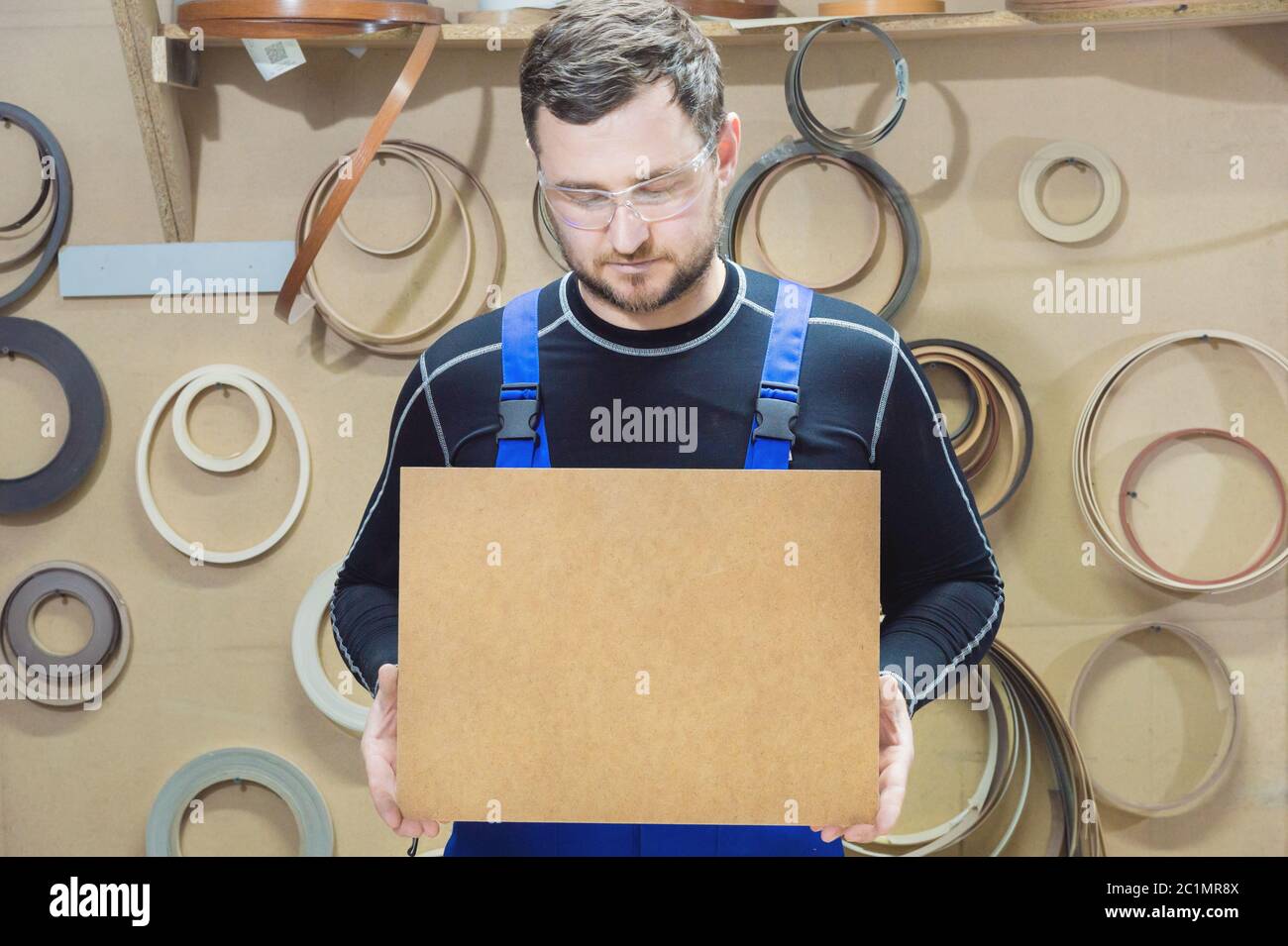 Il produttore di mobili in abiti speciali e occhiali mantiene una piastra vuota per il testo mentre si trova sul posto di lavoro. Imbarcazione p Foto Stock