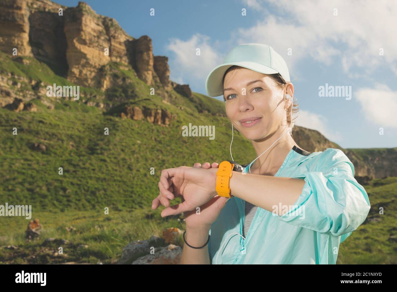 Ritratto di una giovane ragazza sorridente in un cappuccio e cuffie che controlla il suo orologio intelligente mentre si siede all'aperto contro un backg Foto Stock