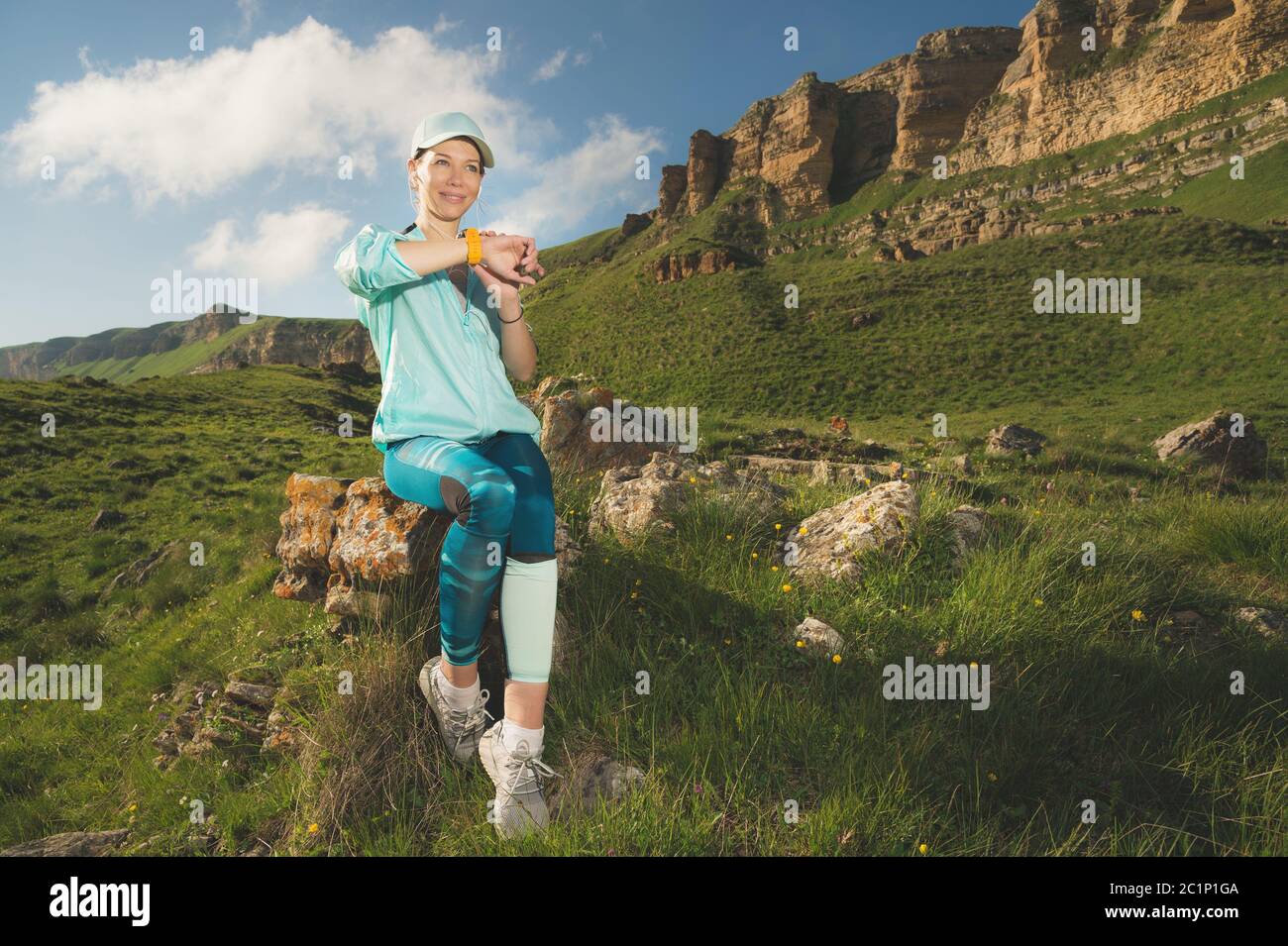 Ritratto di una giovane ragazza sorridente in un cappuccio e cuffie che controlla il suo orologio intelligente mentre si siede all'aperto contro un backg Foto Stock