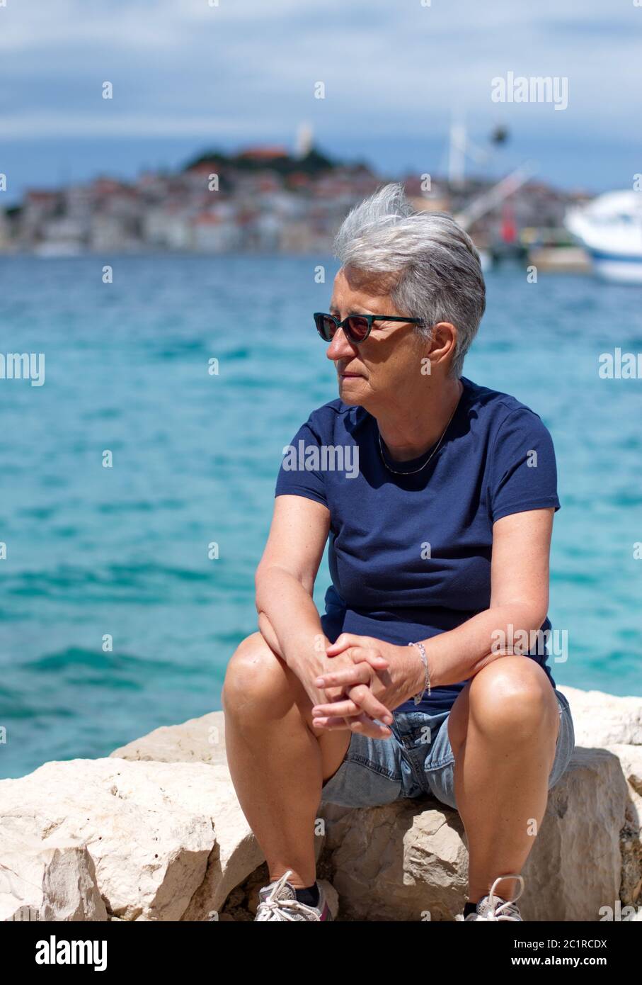 Ritratto di donna anziana seduta sulla riva del mare con città mediterranea sullo sfondo Foto Stock