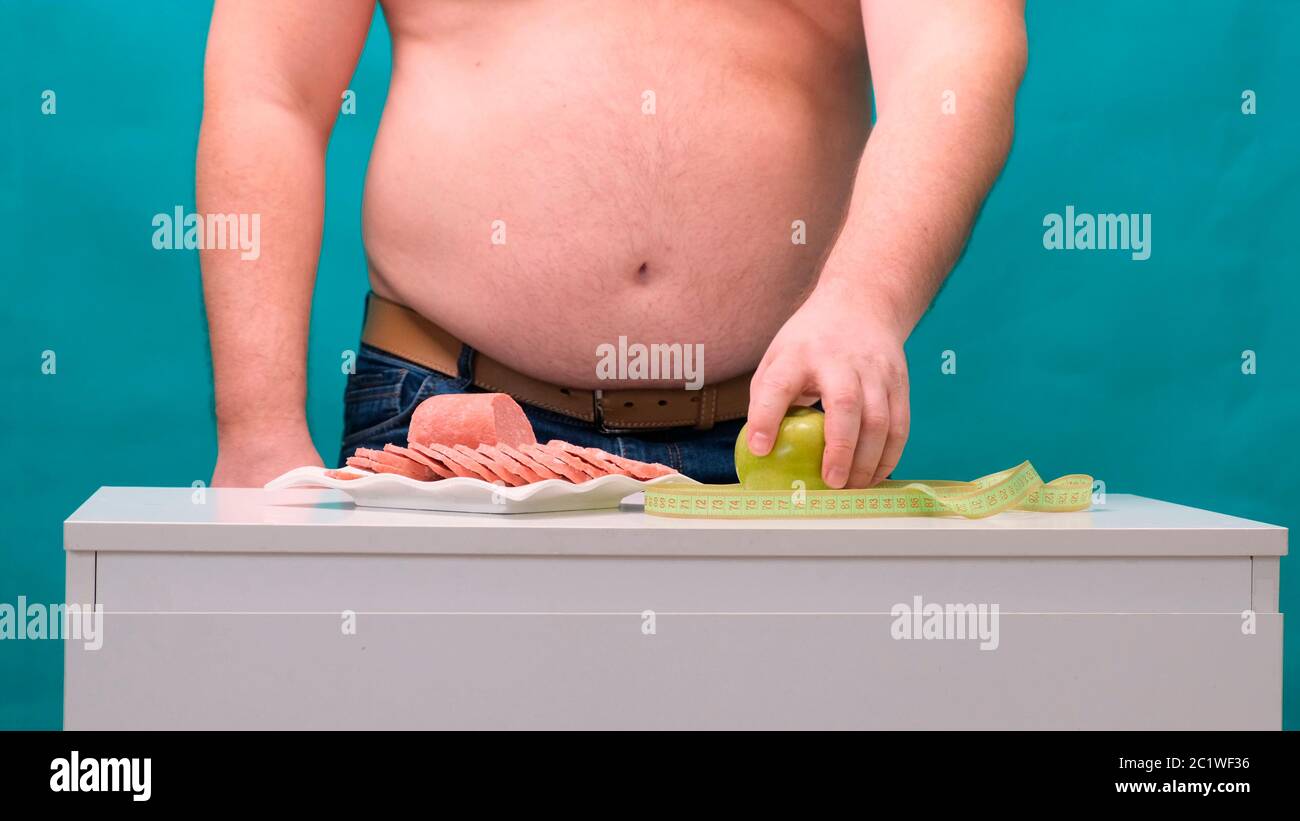 l'uomo con una pancia grassa sceglie tra salsiccia e mela verde. Il concetto di dieta e di volontà. Foto Stock