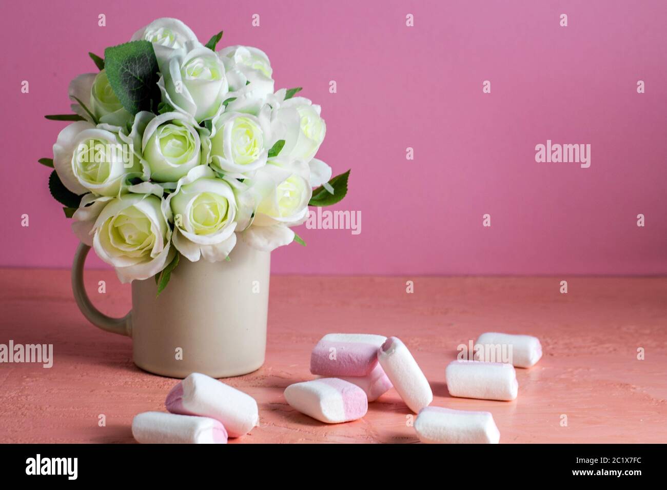 CopySpace con mini marshmallow colorati su un tavolo accanto a rose bianche su sfondo rosa. Foto Stock