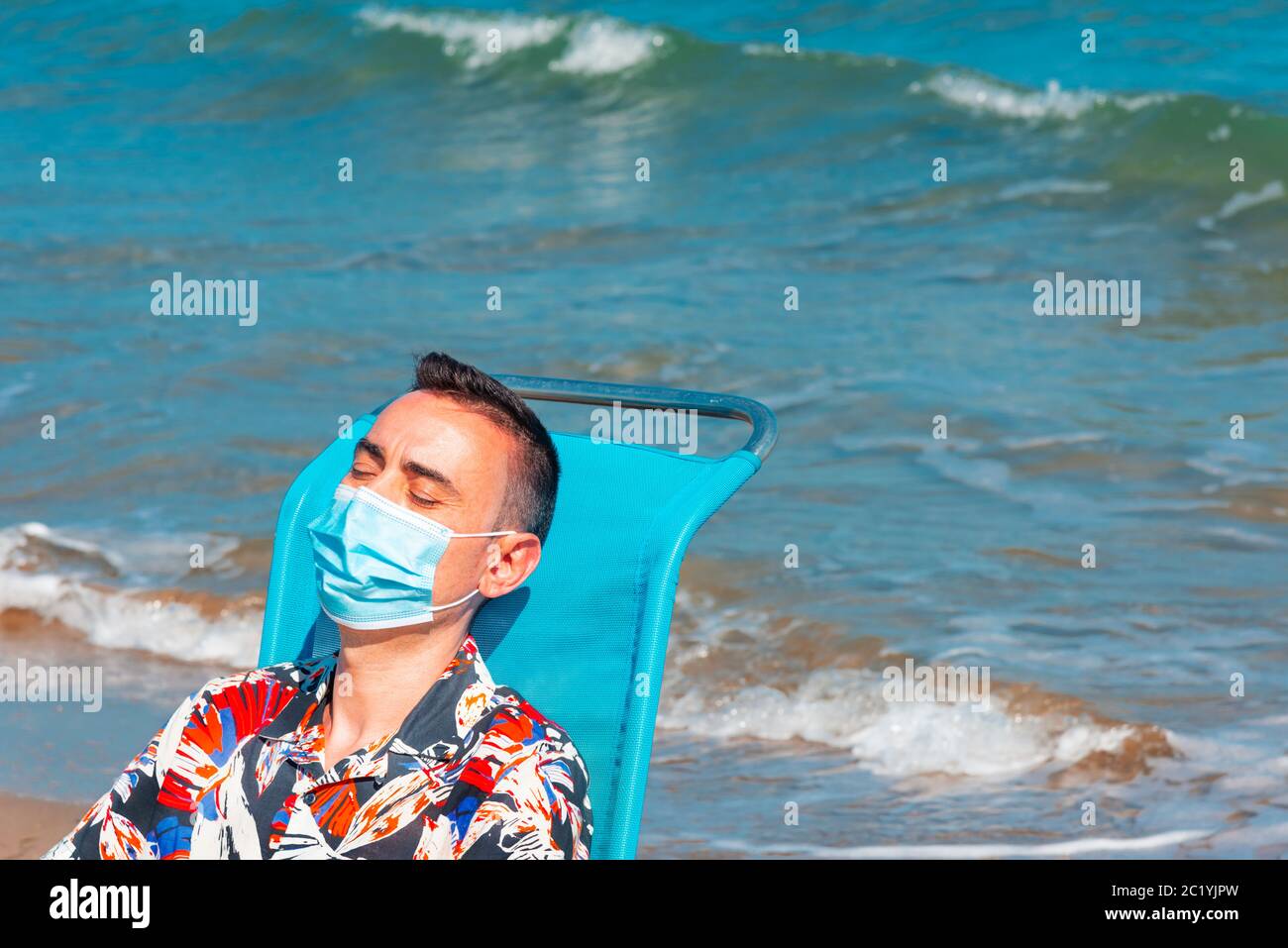primo piano di un uomo caucasico che indossa una maschera chirurgica blu mentre è seduto su una sedia a sdraio sulla spiaggia, con il mare sullo sfondo Foto Stock