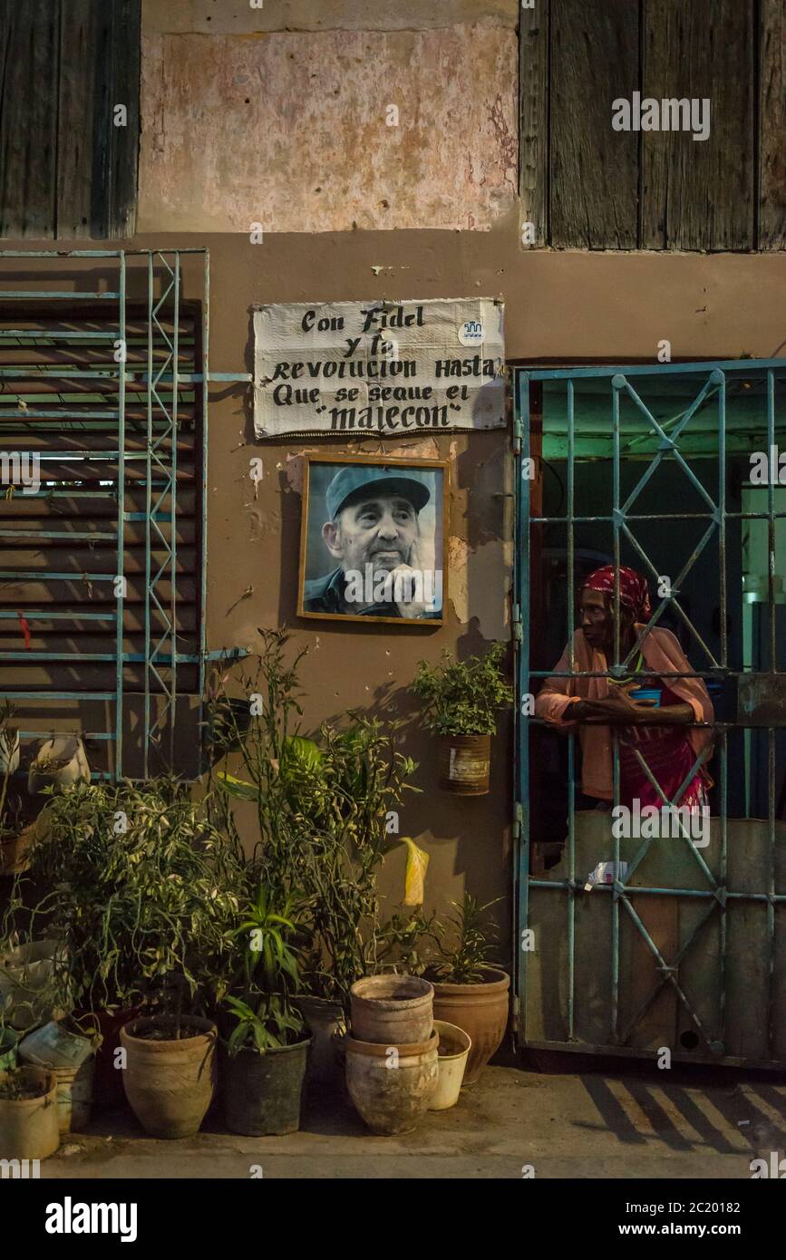 Donna che guarda attraverso i bar della sua porta d'ingresso accanto ad una foto di Fidel Castro a l'Avana Centro, un quartiere di classe operaia, l'Avana, Cuba Foto Stock
