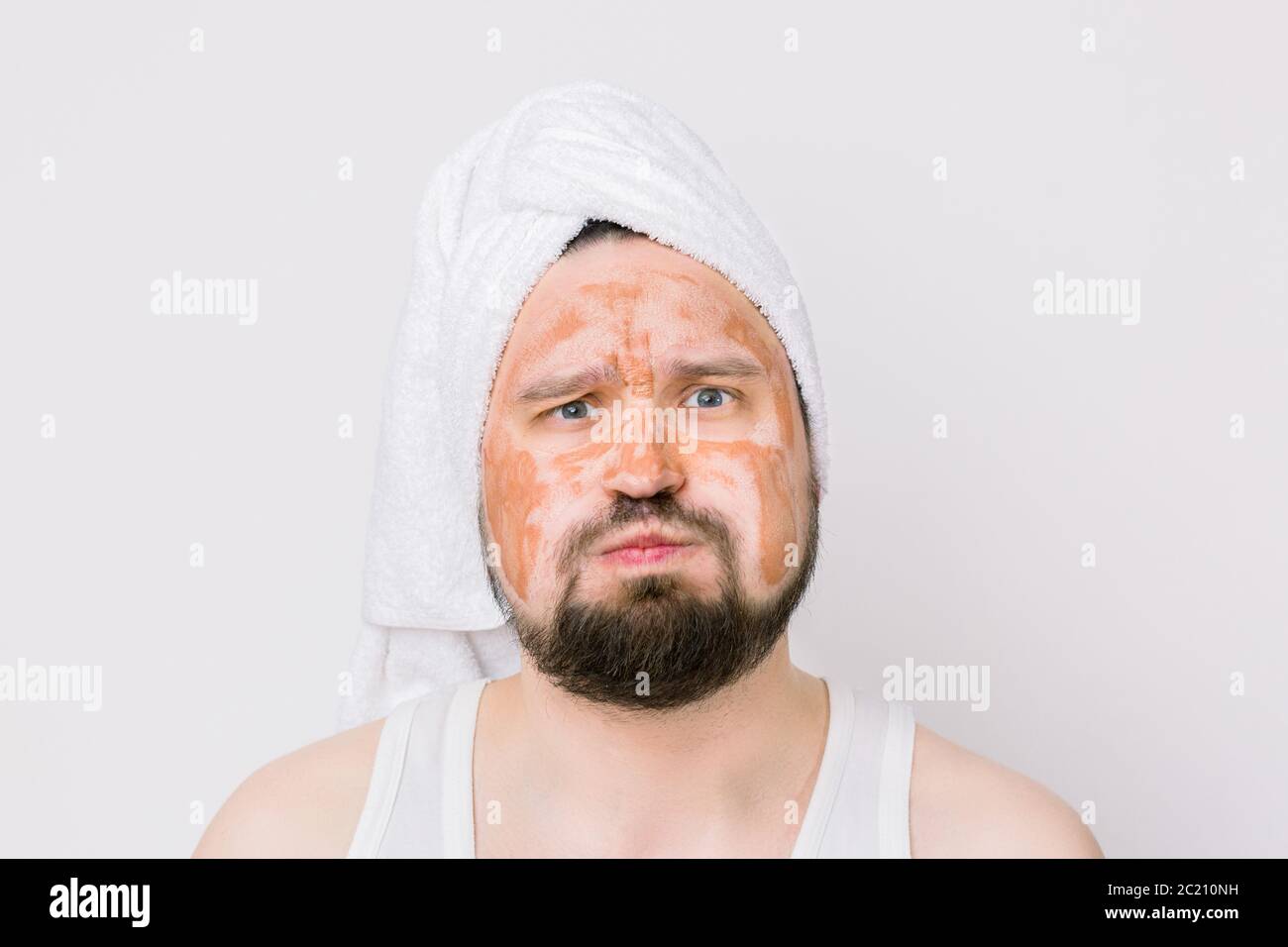 Divertente uomo bearded bello con asciugamano bianco sulla testa, con una maschera marrone fango sul viso, guardando la macchina fotografica su sfondo bianco. Uomo con volto espressivo Foto Stock