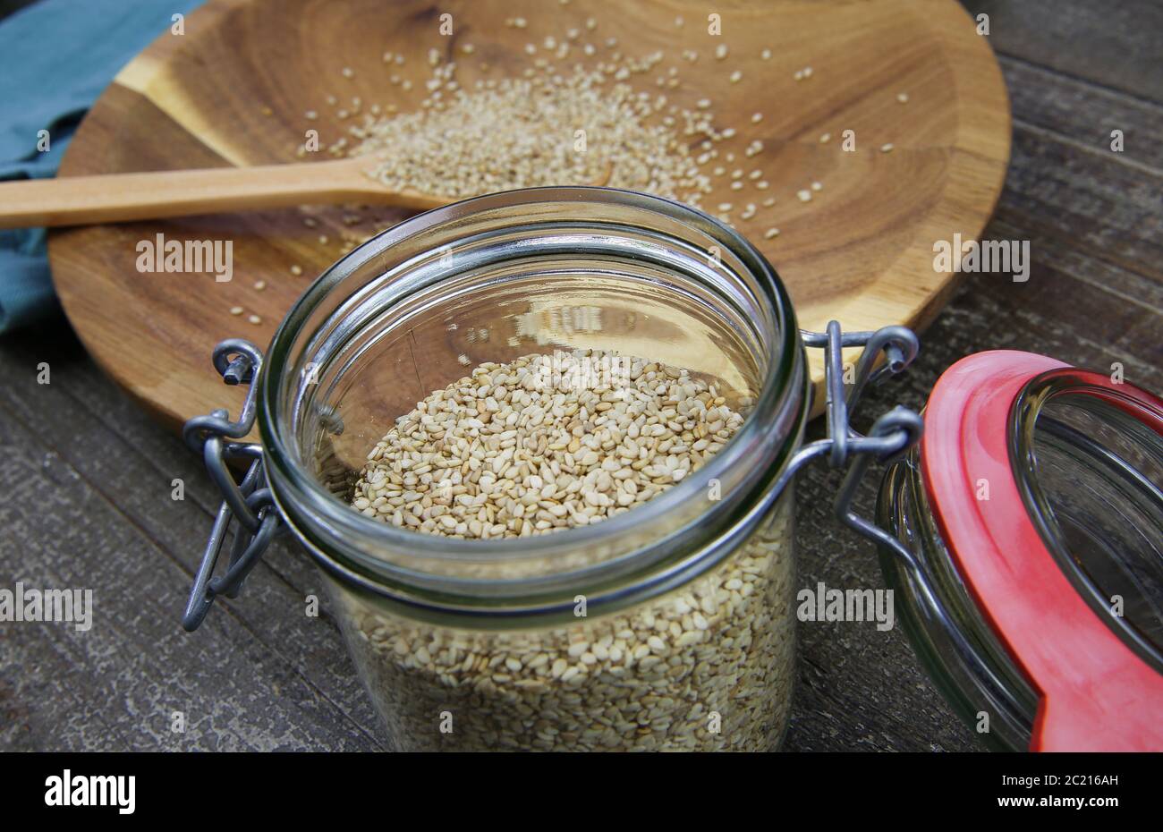 Vista sul vasetto di conservazione con anello in gomma rossa su semi di sesamo grezzi isolati su cucchiaio e piastra in legno Foto Stock