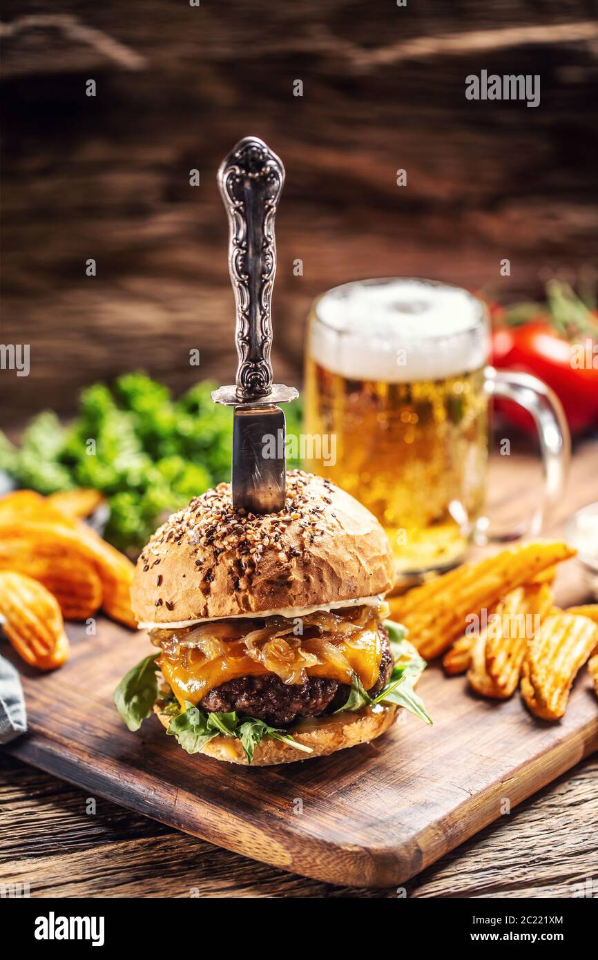 Hamburger di manzo con cipolla caramellata e rucola in un ambiente rustico in legno, una birra e zeppe di patate sul lato Foto Stock