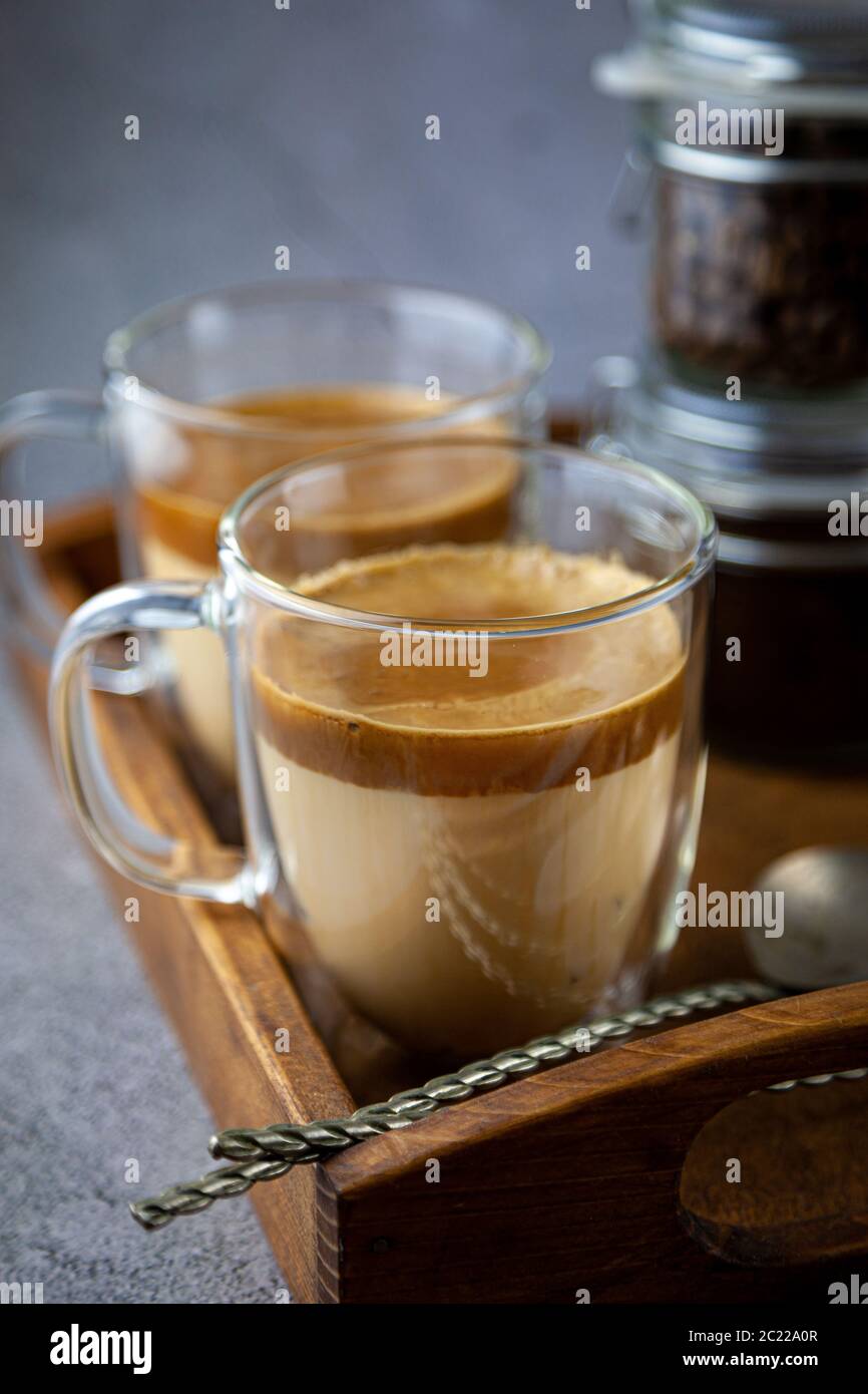 Due tazze trasparenti con pareti doppie. Latte - caffè con latte su un vassoio. La colazione è sul tavolo. Tempo di caffè ancora vivo. Primo piano. Foto Stock