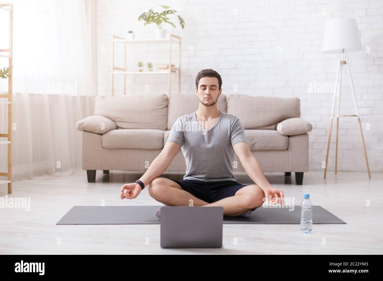 Meditazione e yoga. Un ragazzo con occhi chiusi si siede in posizione lotus sul tappetino, con un laptop Foto Stock