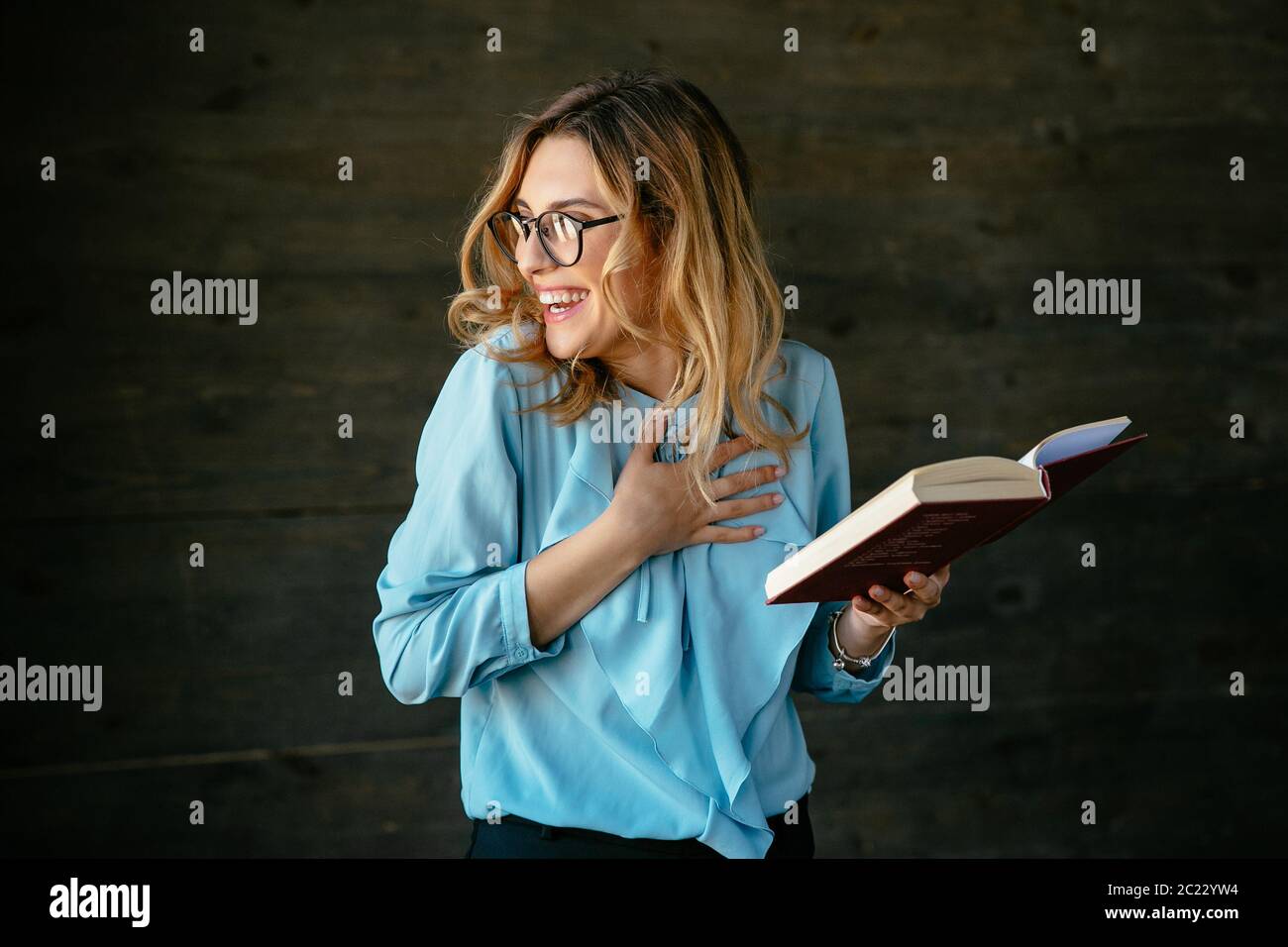 Eccitato giovane donna di ridere dopo aver letto qualcosa di divertente nel libro. Vestito in blu camicetta, in occhiali. Foto Stock