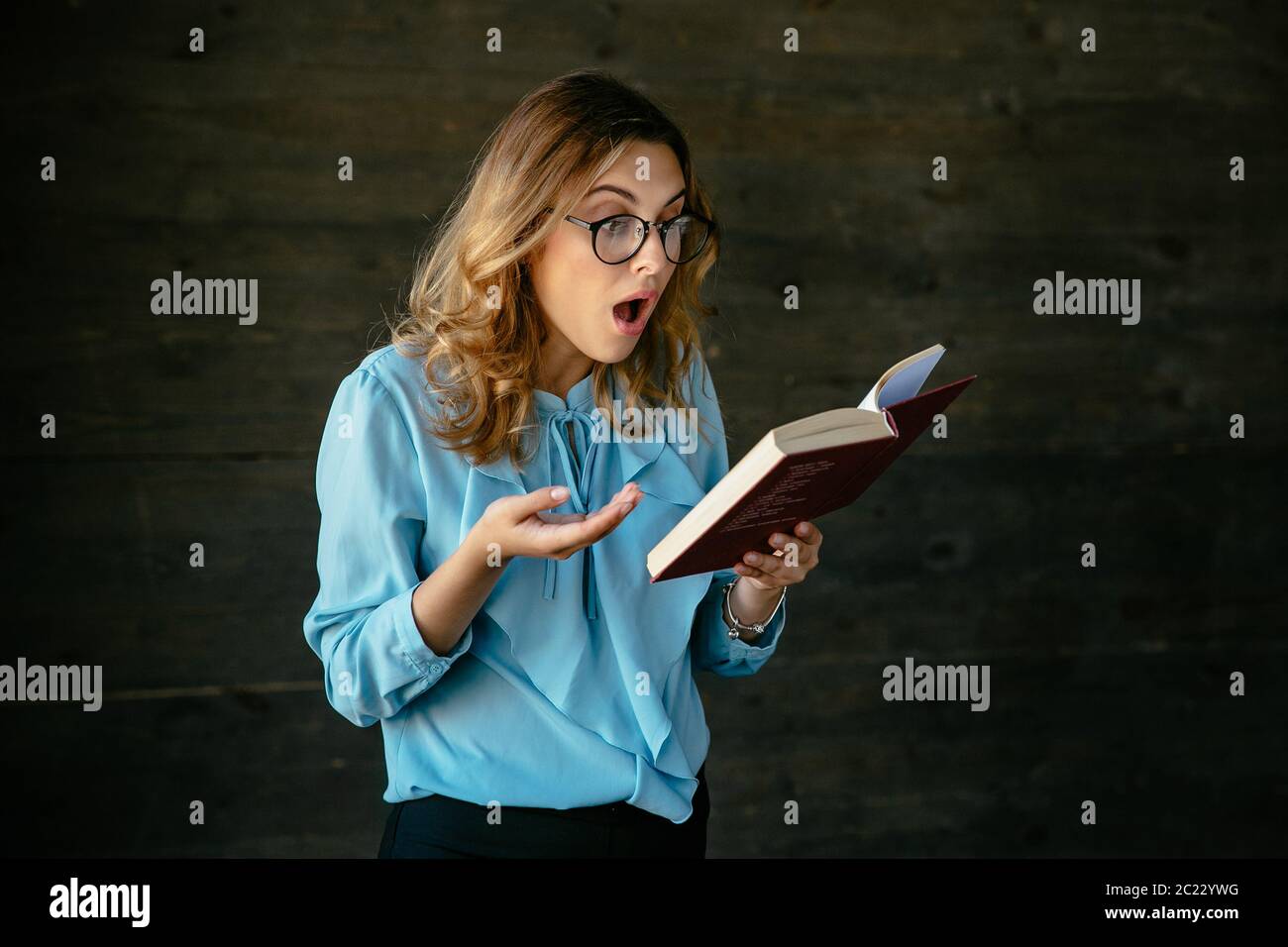 Giovane donna attraente in occhiali sembra sorpreso, con la bocca aperta durante la lettura di un libro. Vestito in Elegante camicetta. Foto Stock