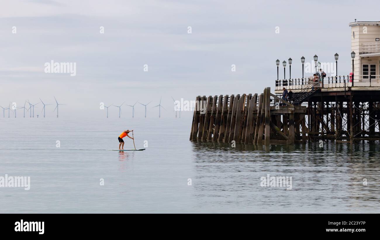 Worthing, Sussex, UK; 15 giugno 2020; Paddleboarder maschio singolo in Bright Orange Fluorescent Top su un mare calmo. Legno Timbers da un molo sulla destra A. Foto Stock
