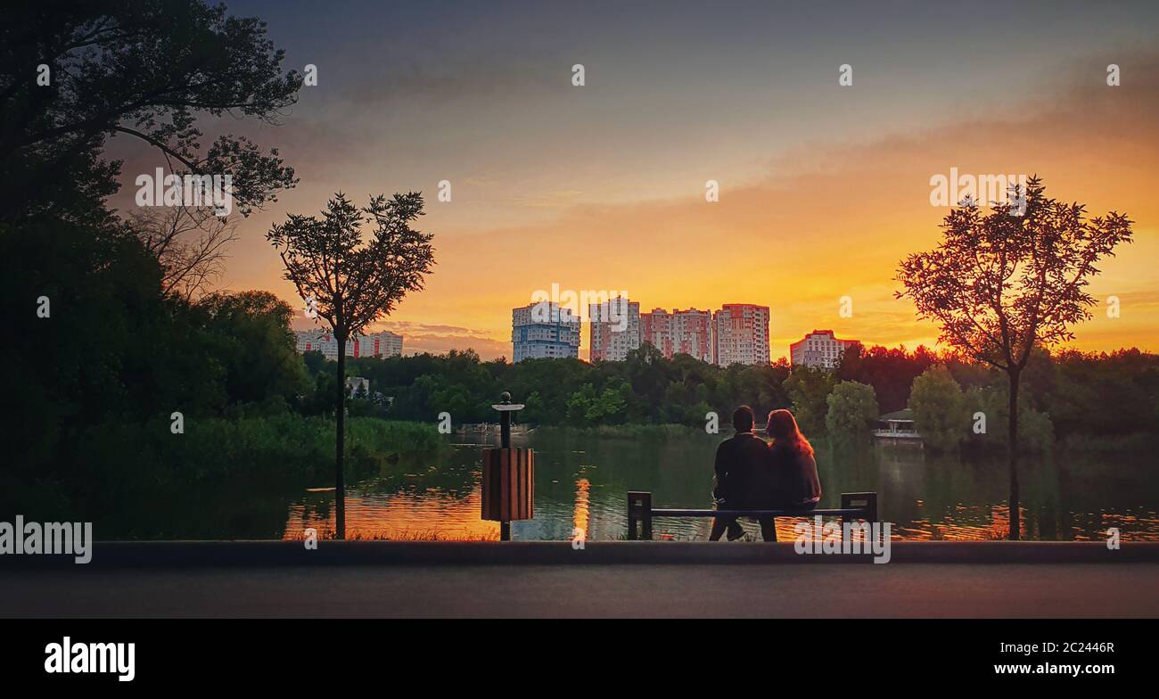 Giovane coppia seduta insieme sulla panchina nel parco vicino al lago contro il tramonto luminoso. Sfondo romantico stile di vita, concetto di togetherness. Amante Foto Stock