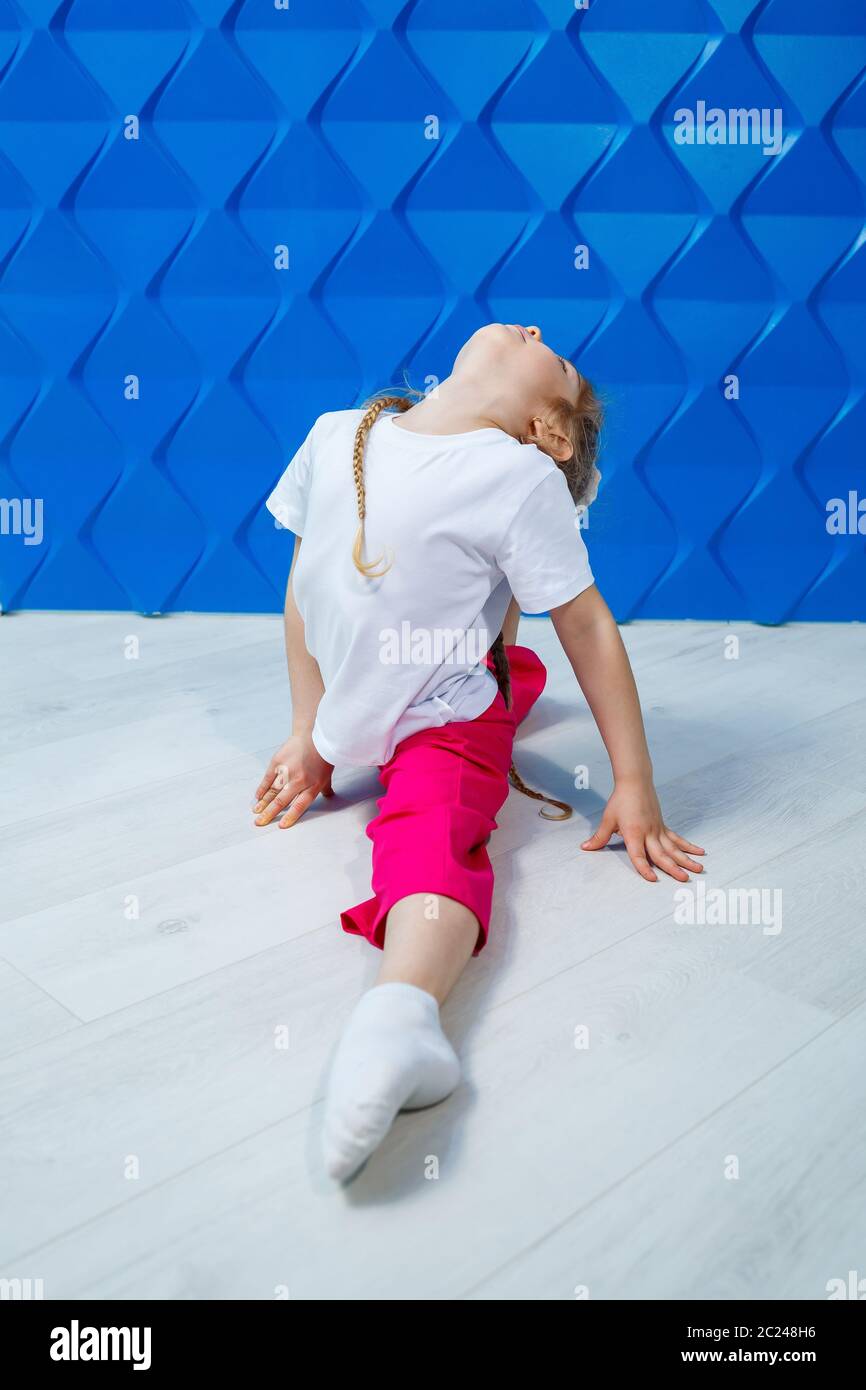 Una bambina con i pigtail in una T-shirt bianca su uno sfondo blu si siede in spago sul pavimento e sorride. Emozioni dei bambini occupazione spot, divertimento Foto Stock