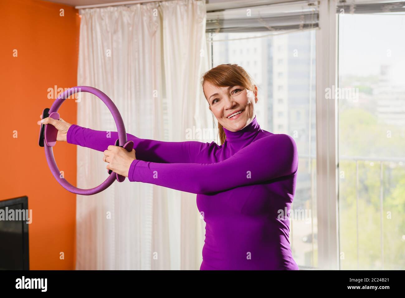 Ritratto di sorridente positivo sportivo donna di mezza età fare ginnastica mattina, allenarsi a casa le mani durante l'isolamento con strumento circolare ad anello Foto Stock