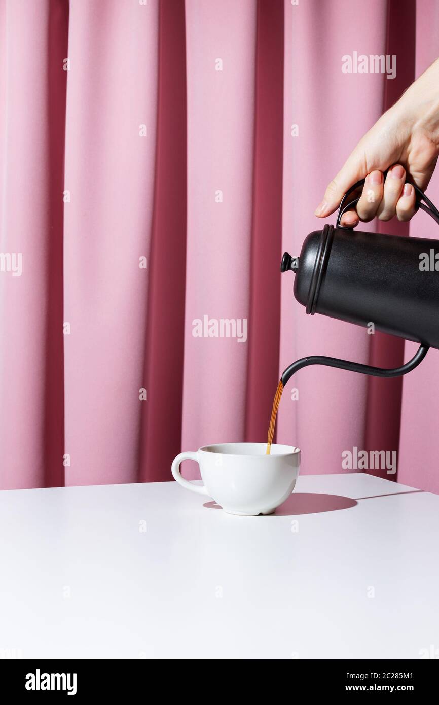 Mano della donna che tiene il vaso di metallo nero e che versa il caffè o il tè in una tazza bianca davanti al tendaggio rosa, fuoco selettivo Foto Stock