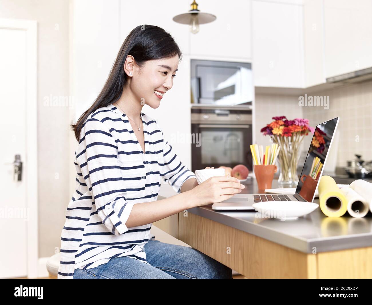 giovane donna d'affari asiatica che lavora da casa seduto al banco della cucina che tiene una tazza di caffè guardando il computer portatile, felice e sorridente Foto Stock