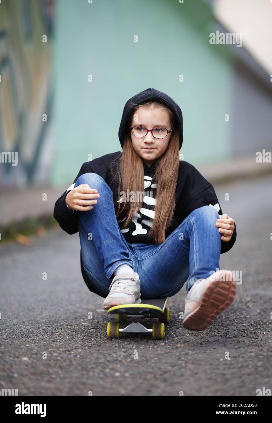 La ragazza corse su skateboard. Bambino in una felpa nera seduta sul suo skateboard. Colpo verticale. Messa a fuoco selettiva. Foto Stock