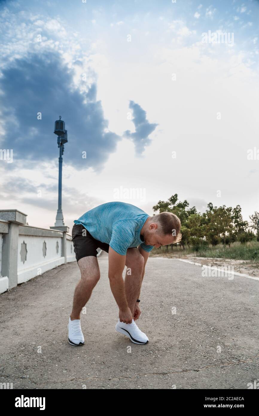 Ritratto di ragazzo caucasico in una maglietta blu e pantaloncini nero,che lega il suo laces su bianco calzature sportive prima di jogging Foto Stock