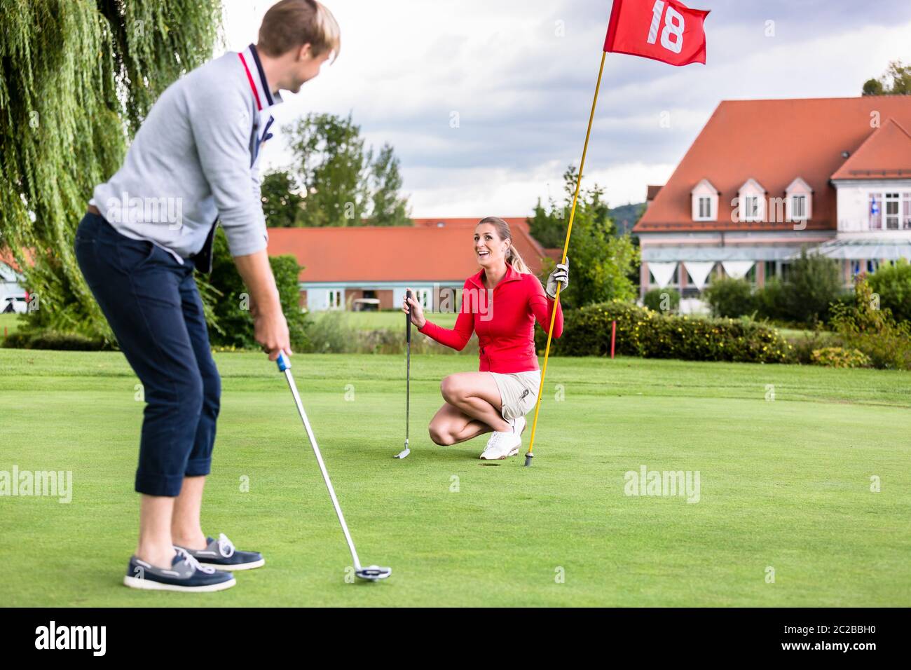 Sorridente ragazza tenendo bandiera guardando golfista maschio di mettere la pallina da golf nel verde Foto Stock