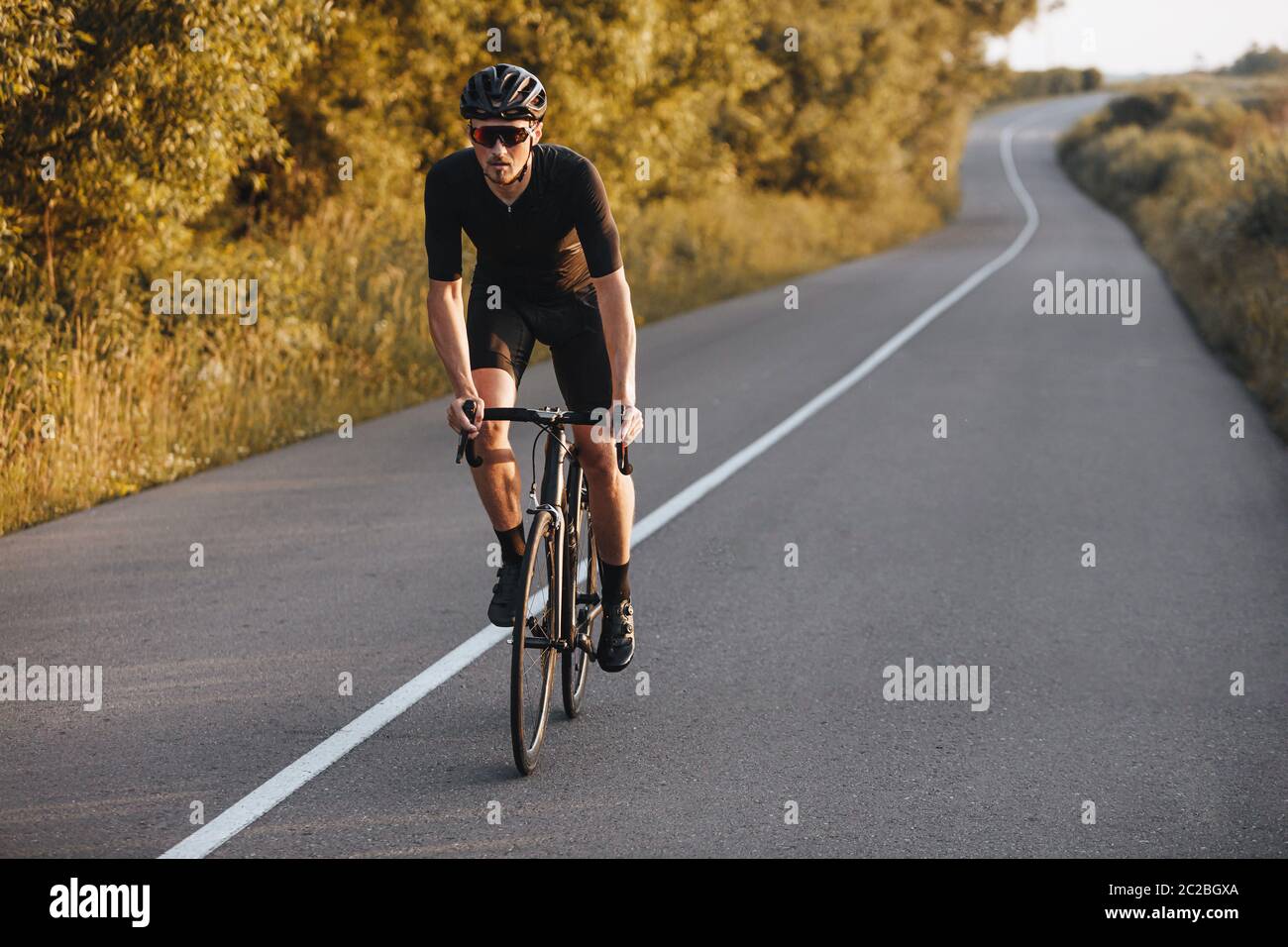 Atleta forte in bici vestiti equitazione su strada asfaltata con alberi verdi intorno. Ciclista attiva in casco nero e occhiali a specchio facendo Foto Stock