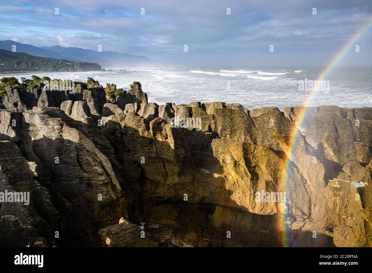 Un arcobaleno è formato da spray marino nell'aria dopo un'eruzione di grandi fioriture a Punakaiki sulla costa occidentale dell'isola meridionale della Nuova Zelanda. Foto Stock