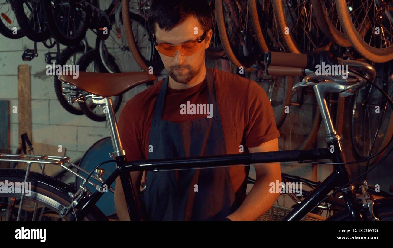 Tema piccola impresa bici riparazione. Giovane uomo caucasico bruna indossare occhiali di sicurezza, guanti e grembiule usa la riparazione di attrezzi manuali Foto Stock