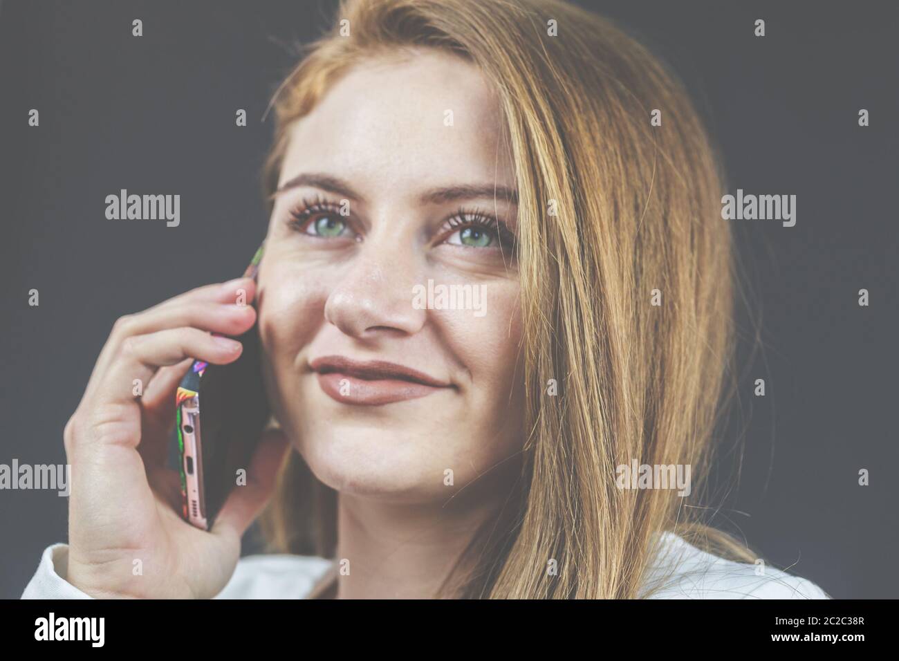 Giovane donna con jeans e blazer bianco sorride e tiene il suo smartphone in mano, telefonando e leggendo notizie. Foto Stock