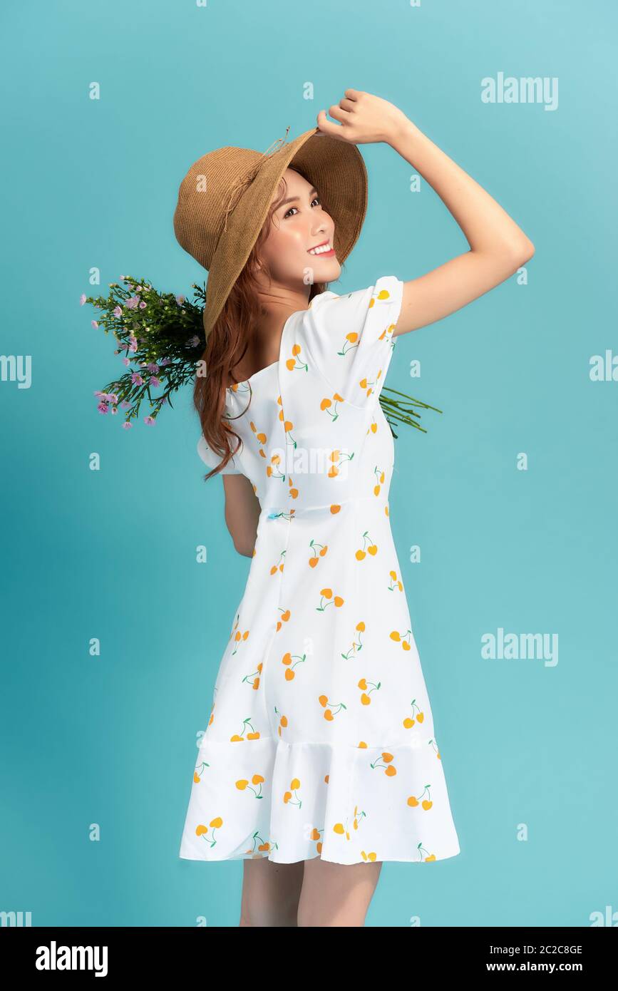 Attraente giovane donna in abito estivo e cappello di paglia che tiene bouquet margherita e guardando sopra la sua spalla isolata su sfondo blu Foto Stock