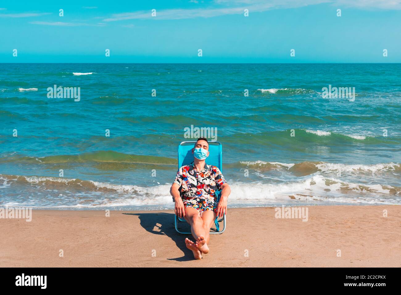 un uomo caucasico che indossa una maschera chirurgica blu mentre è seduto su una sedia a sdraio sulla spiaggia, con il mare sullo sfondo Foto Stock