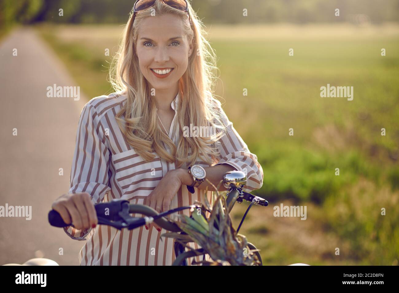 Donna che usa la sua bicicletta per acquistare prodotti freschi in piedi su una strada rurale retroilluminato da un caldo bagliore del sole sorridendo felice alla macchina fotografica come la tiene Foto Stock