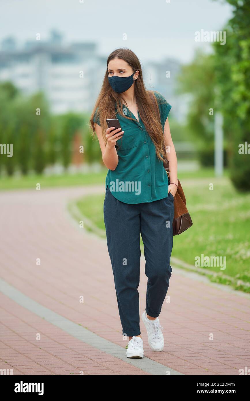 Donna in maschera medica legge notizie su smartphone che tinte mano nella tasca dei pantaloni passeggiate nel parco. Ragazza che mantiene la distanza sociale Foto Stock