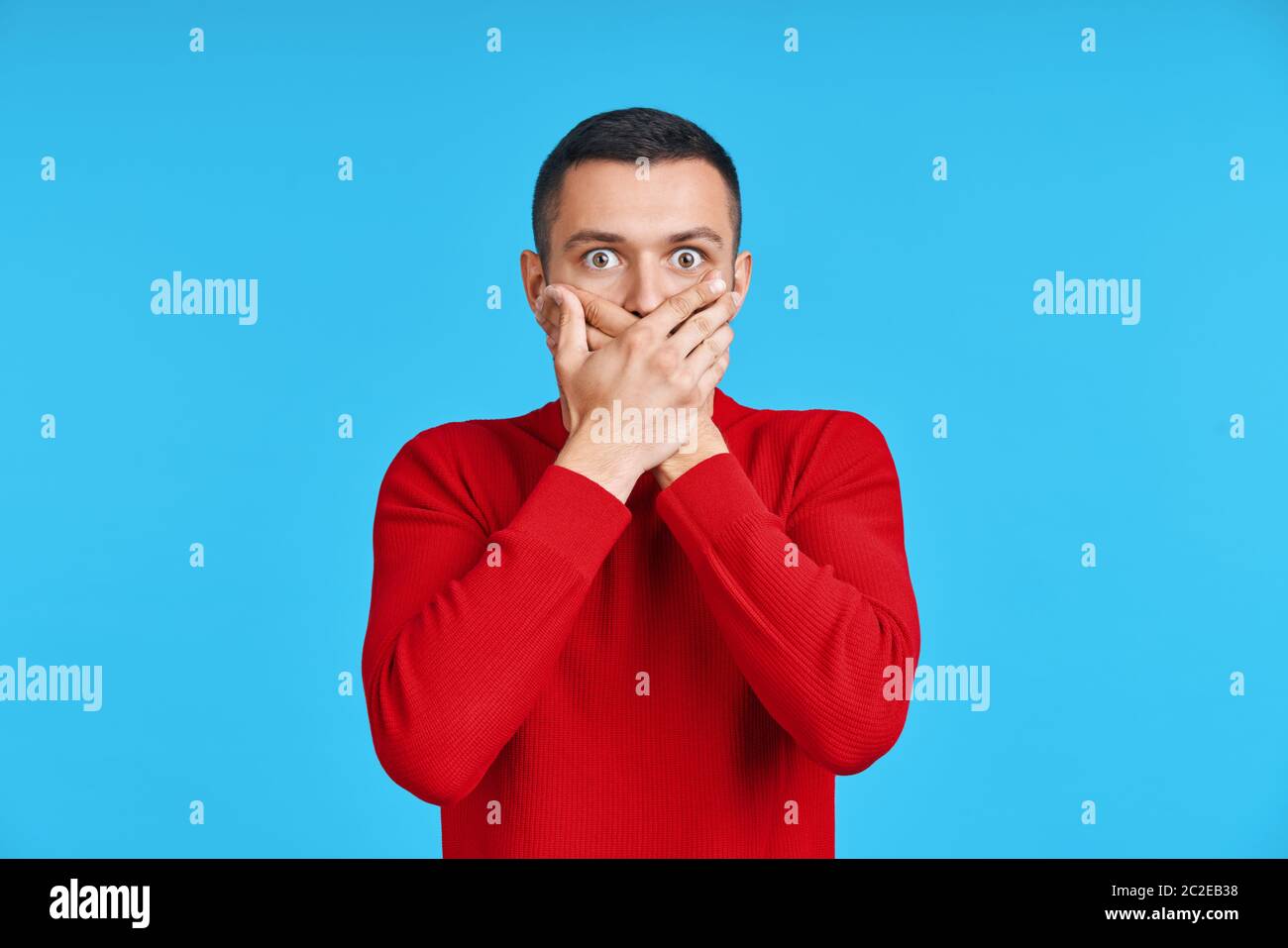 Uomo scioccato che copre la bocca con le mani su sfondo blu Foto Stock