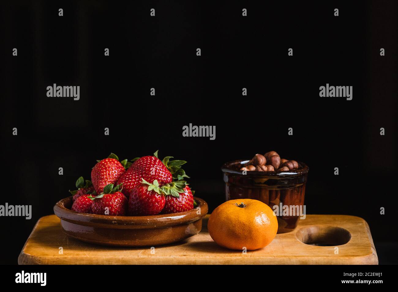Ancora vita con fragole, nocciole e un mandarino con contenitori in ceramica su una tavola di legno da cucina Foto Stock