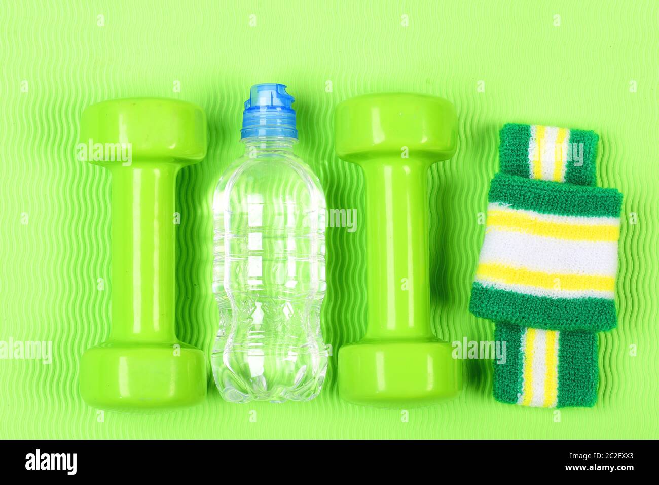 Coppia di manubri verdi per fitness e bottiglia d'acqua su sfondo verde. Due manubri in plastica, braccialetti e bottiglia. Sport, dieta e perdita di peso concetto. Set di pesi manuali su sfondo luminoso. Foto Stock