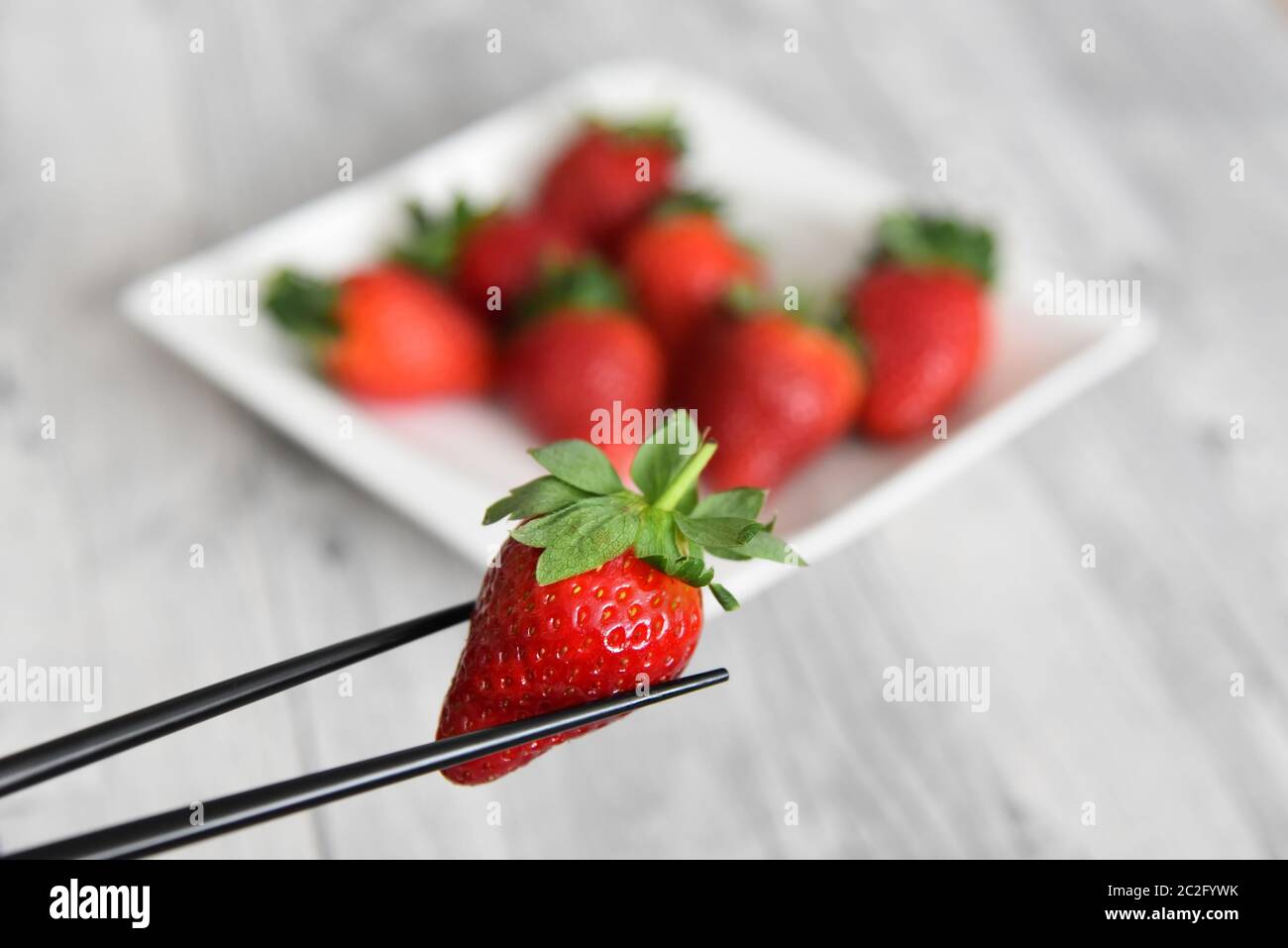 Bastoncini di pollo che tengono una fragola rossa fresca davanti a uno sfondo sfocato di un piatto di fragole. Foto Stock