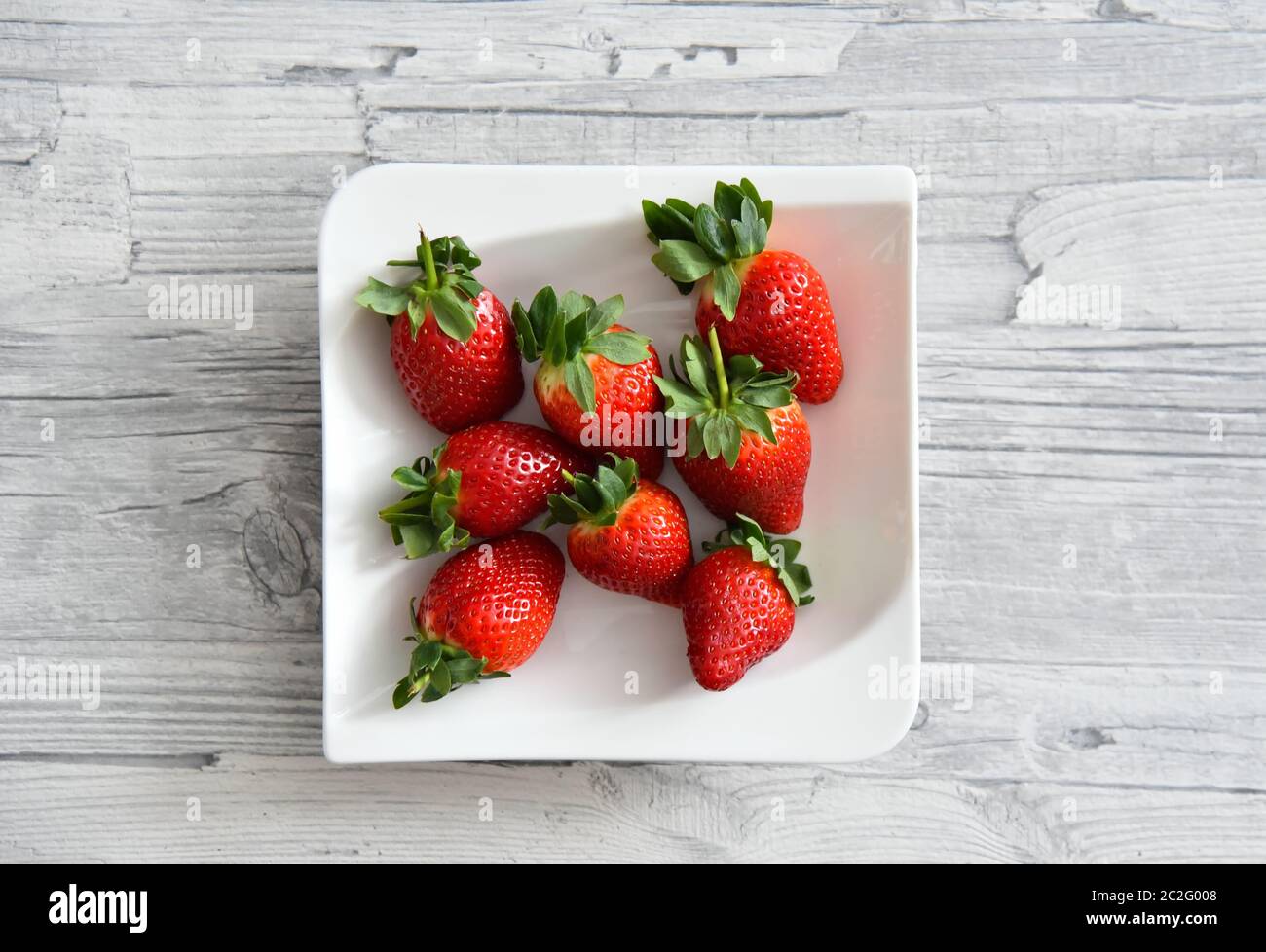 Fragole rosse fresche e succose con foglie verdi adagiate su un piatto bianco. Sfondo tavolo in legno grigio chiaro. Foto Stock