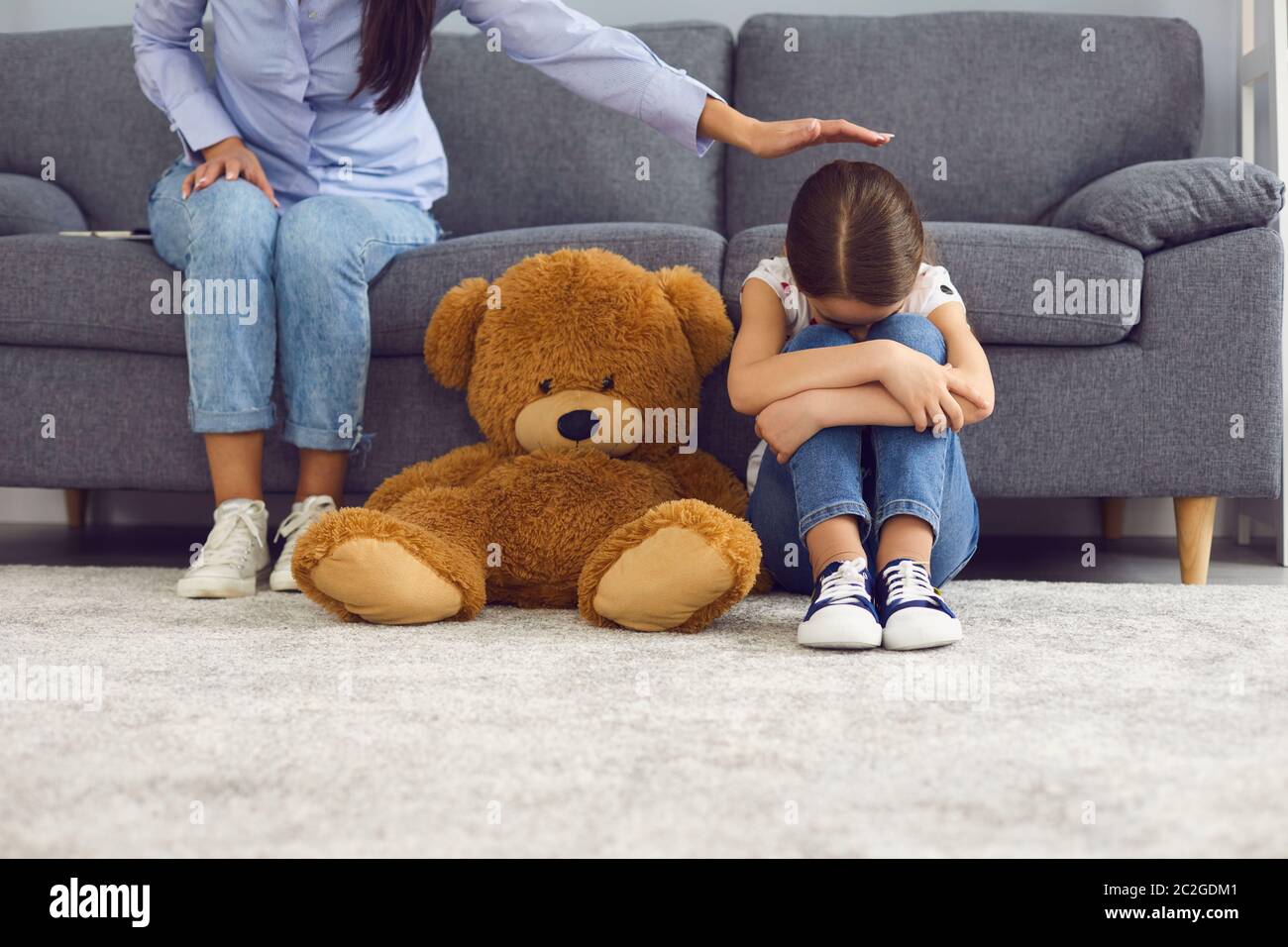 La bambina sconvolta piangendo sul pavimento vicino orsacchiotto mentre la madre la conforta a casa. Genitore calmante il suo bambino infelice Foto Stock