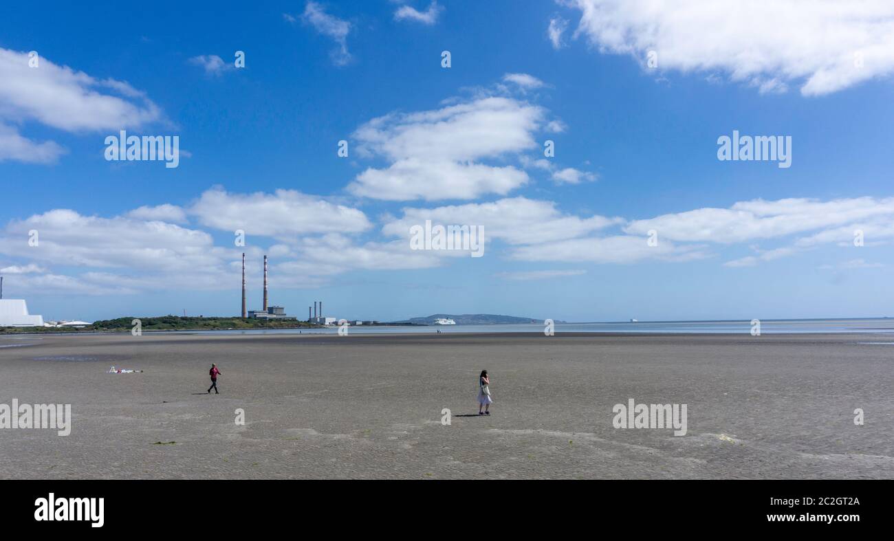 La gente cammina lungo Sandymount Strand a Dublino con le torri gemelle di Poolbeg in lontananza e un traghetto che entra nel porto di Dublino. Foto Stock