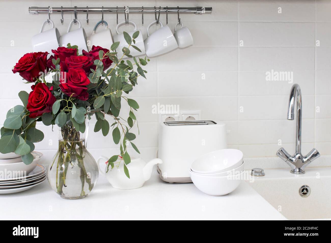 bouquet di rose rosse e eucalipto in vaso di vetro su tavolo da cucina. Tè nero in una tazza bianca, vaso di miele e cucchiaio per miele, primo piano di limone su a. Foto Stock