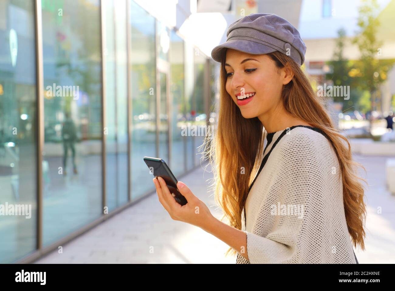 Sorridenti moda donna con baker Boy cappello a piedi in strada e ricevere buone notizie su il suo smart phone. Foto Stock