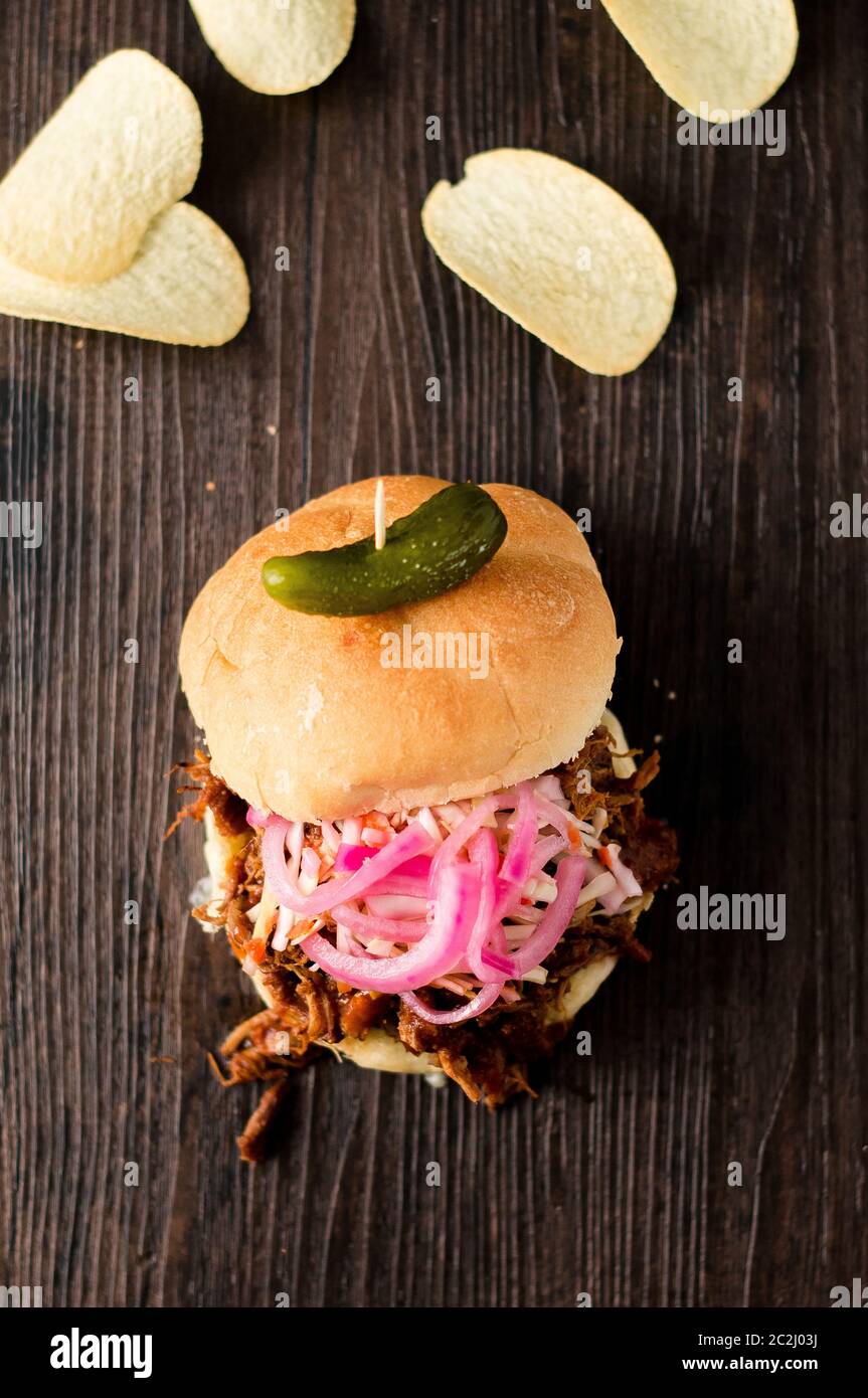 Carne di maiale tirata a cottura lenta con salsa barbecue, cipolle sottaceto, insalata di cavolo e formaggio serviti in un panino hamburger con sottaceto di aneto e patatine come lati. È un caos Foto Stock