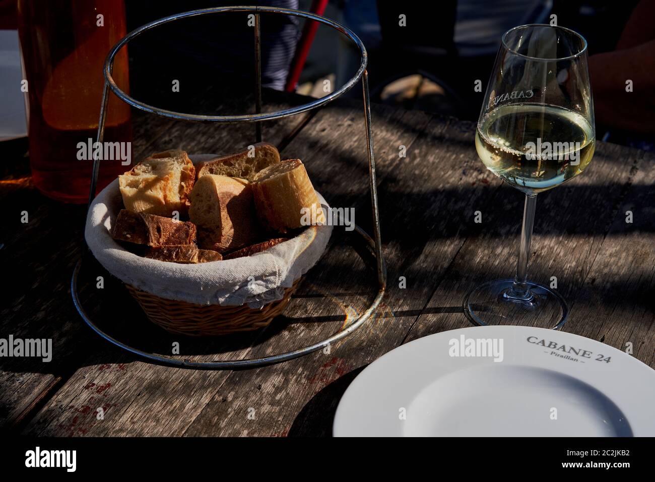 Un cestino di pezzi di baguette e un bicchiere di vino bianco, il bicchiere e il piatto con la scritta Cabane 24, su un tavolo di legno illuminato dal sole, Piraillan Foto Stock