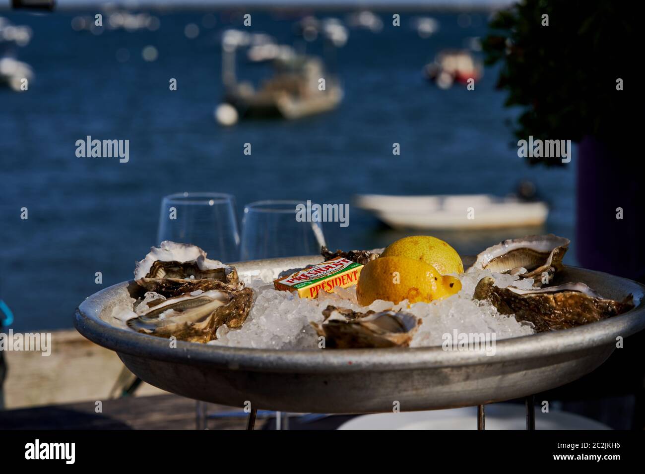 Baia di Arcachon con pescherecci e imbarcazioni a vela sullo sfondo con ostriche servite su un letto di ghiaccio con burro, limone e bicchieri da vino sul davanti Foto Stock